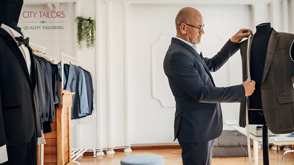 Tailor adjusting a bespoke suit on a mannequin inside City Tailors studio with the City Tailors logo displayed.